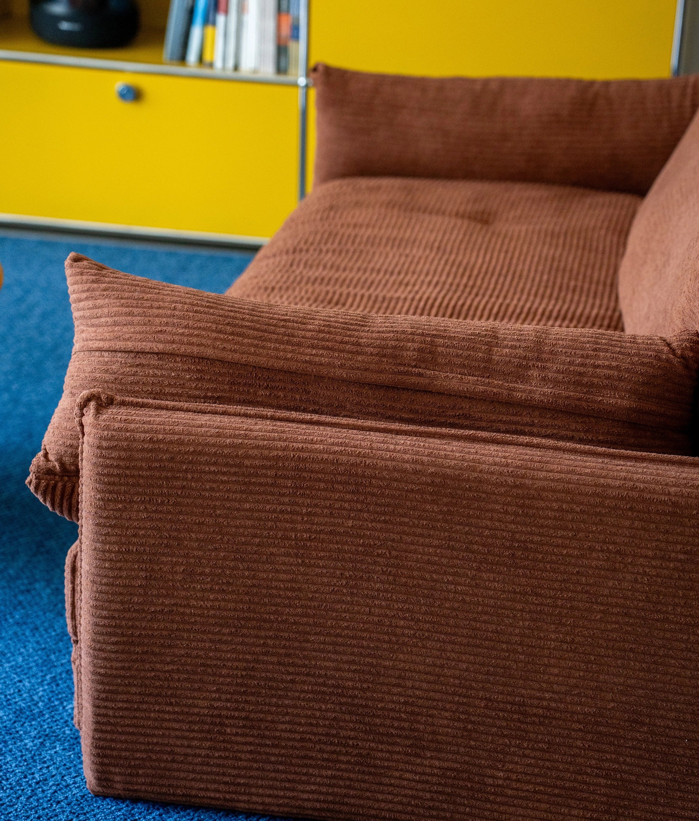 Brown corduroy sofa against a yellow wall with books on a shelf.